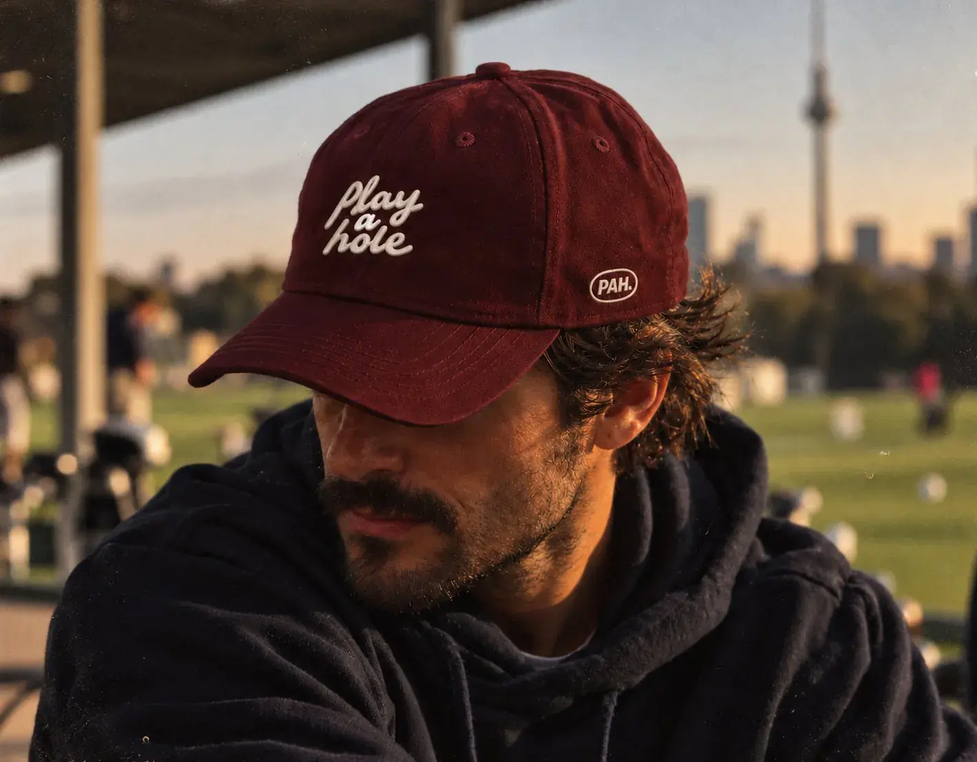 Mann im dunklen Hoodie mit weinroter Play-a-hole-Cap auf der Driving Range, im Abendlicht.