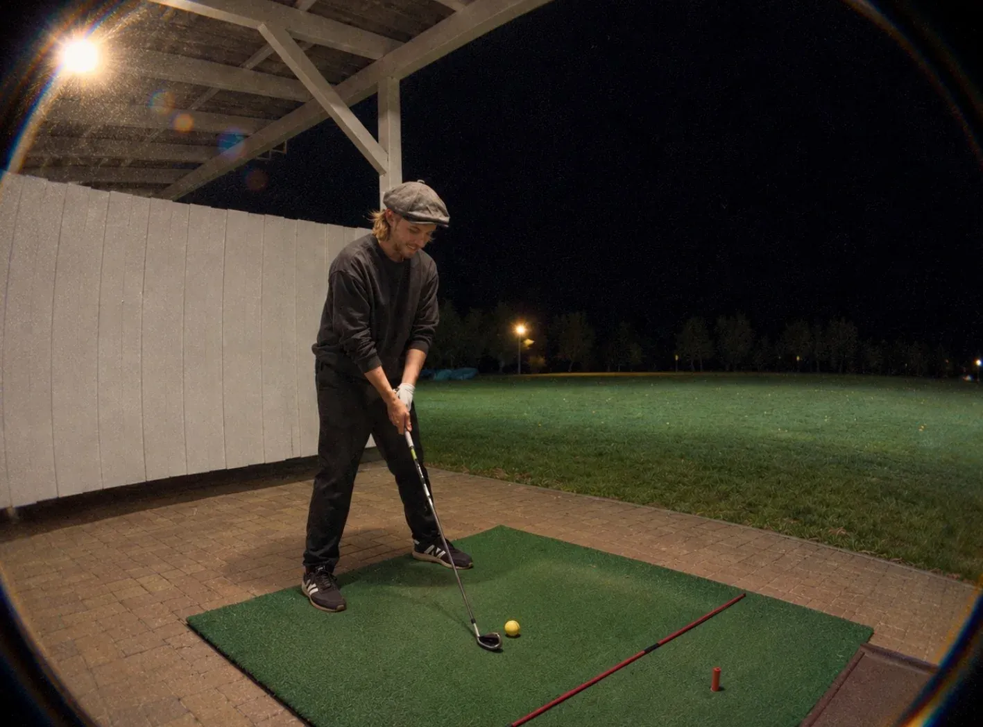 Golfer mit Schiebermütze schlägt abends unter Flutlicht auf der Driving Range ab.