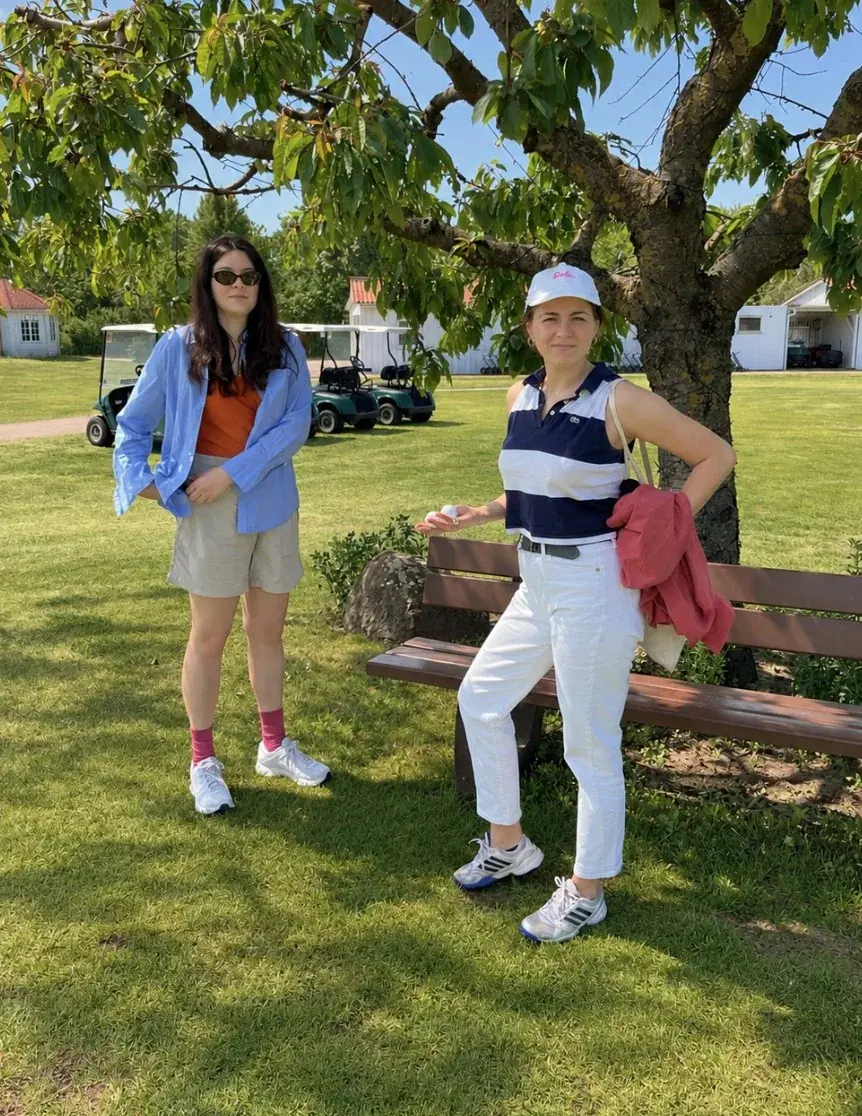 Zwei Golferinnen unter einem Baum neben einer Bank, im Hintergrund Golfcarts.