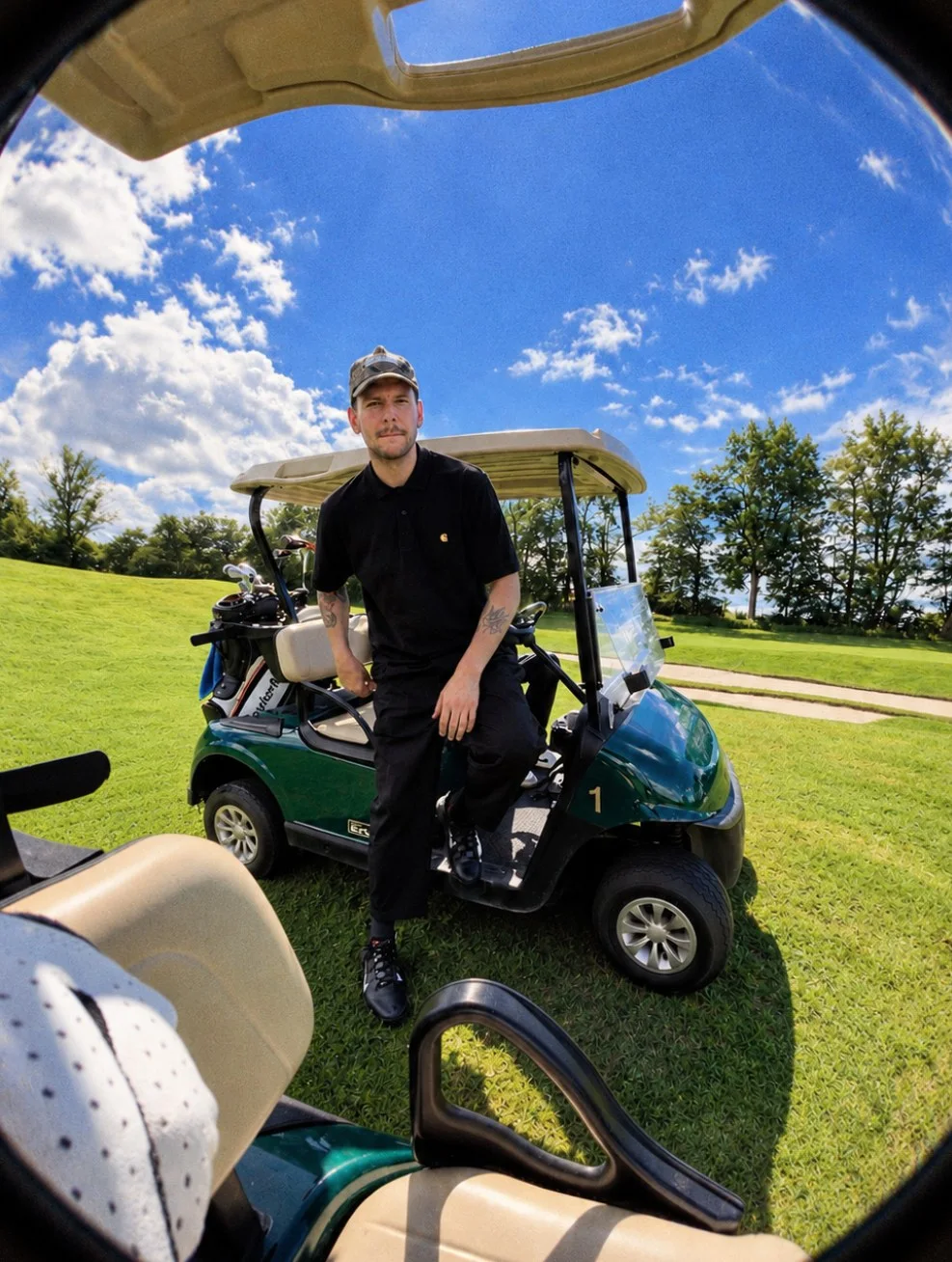 Golfer mit Cap lehnt am Golfcart, mit Fischauge aus einem zweiten Cart fotografiert.