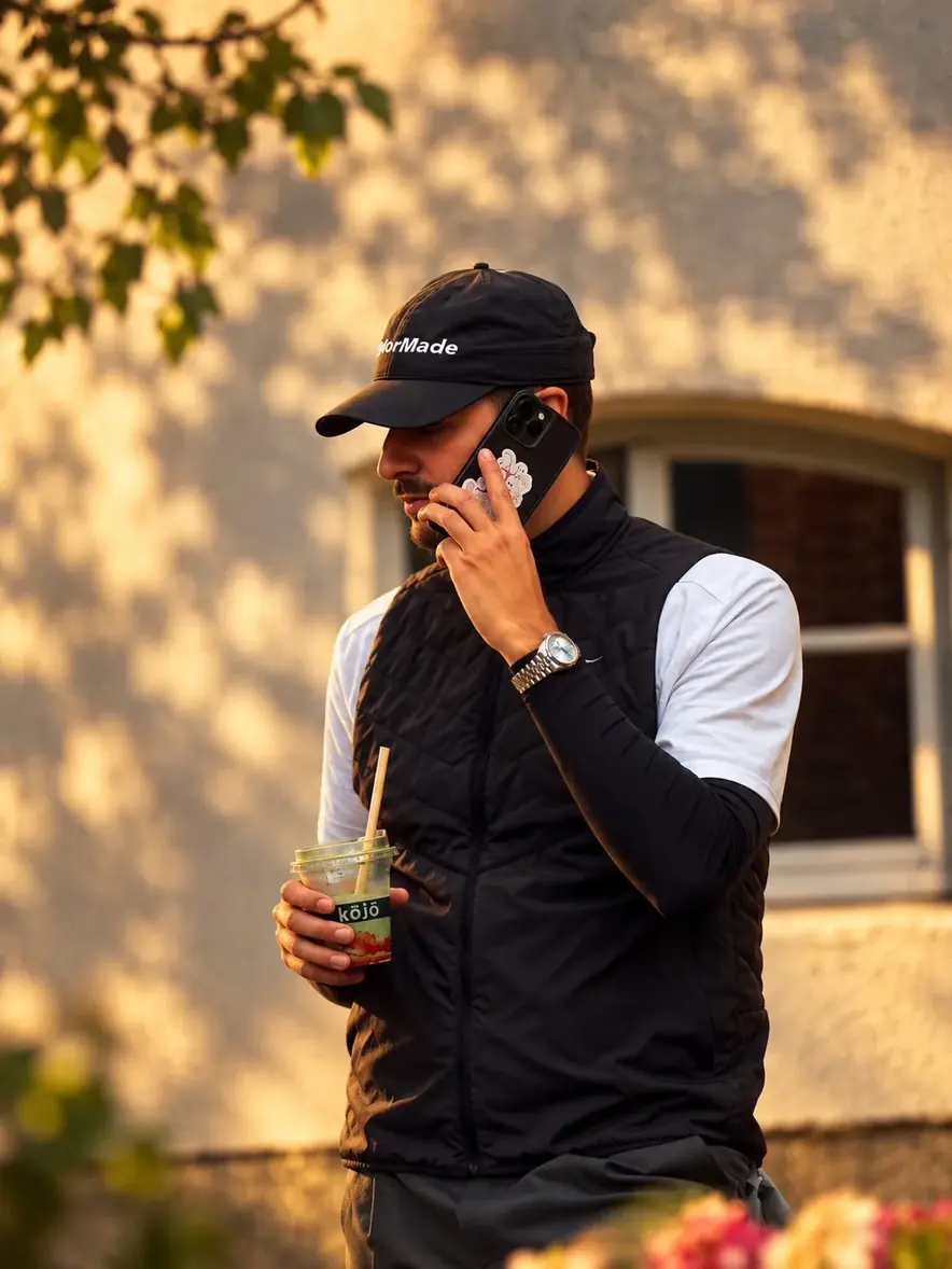 Golfer mit Cap telefoniert im Abendlicht, Drink in der Hand.