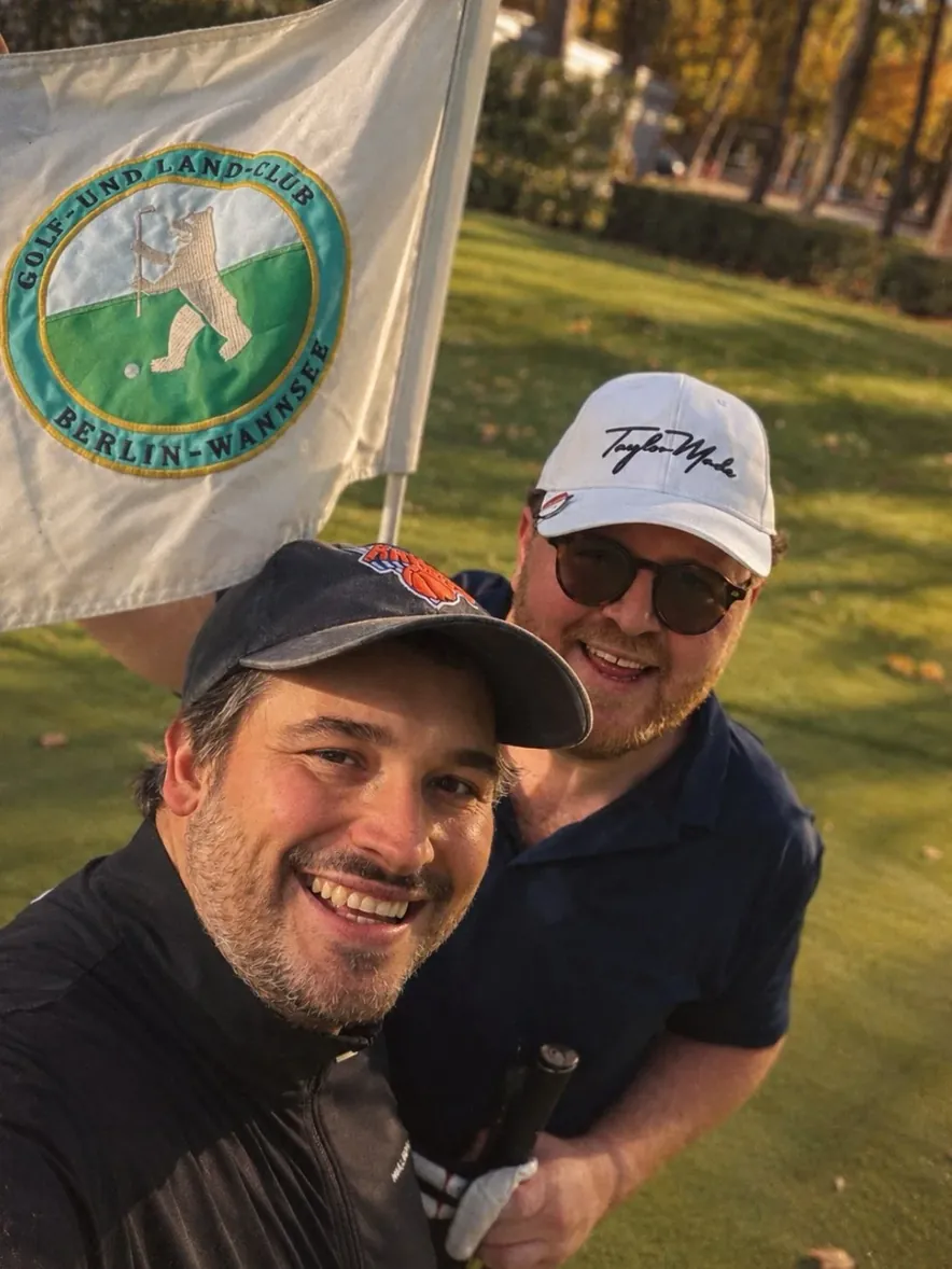 Selfie von zwei lachenden Golfern mit der Fahne vom Golf- und Land-Club Berlin-Wannsee.